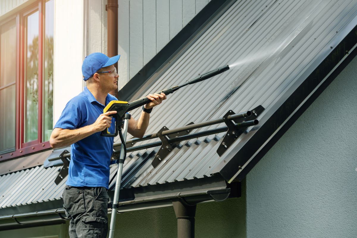 A person in a blue shirt uses a pressure washer to clean a metal roof, with sunlight reflecting off the surface and nearby windows.