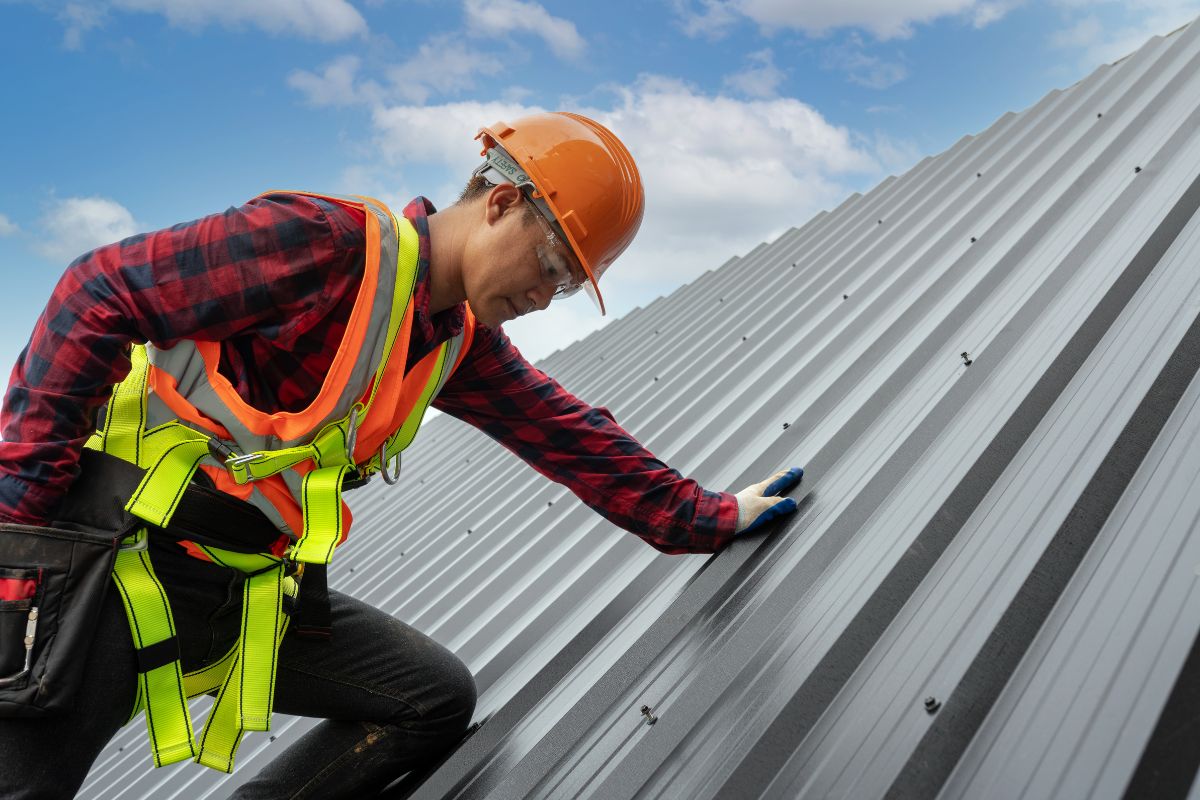 A construction worker in safety gear inspects a metal roof under a bright blue sky with scattered clouds.