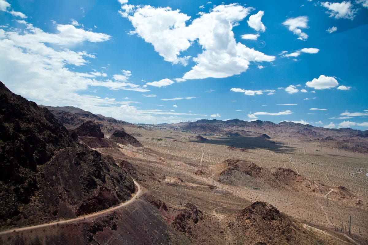 Vast desert landscape under a blue sky, featuring rugged mountains, winding dirt roads, and scattered clouds. A scenic and arid view.