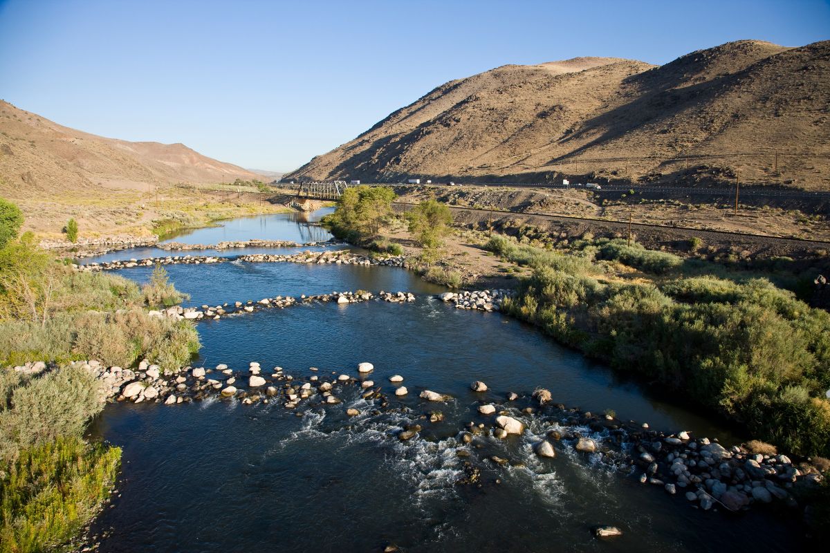 A serene river winds through rocky terrain and hills under a clear blue sky, with lush green vegetation along the banks.