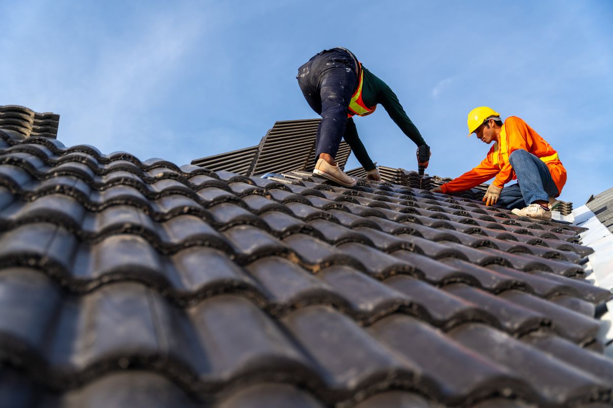 Two men are repairing a roof under a clear blue sky, focused on their work and collaborating effectively.